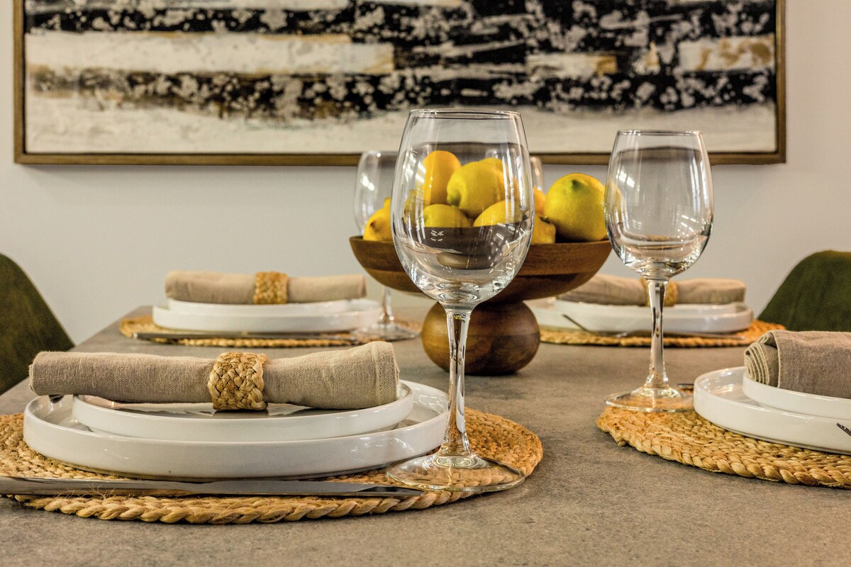 A dining table set with white plates, clear wine glasses, and a bowl of lemons.