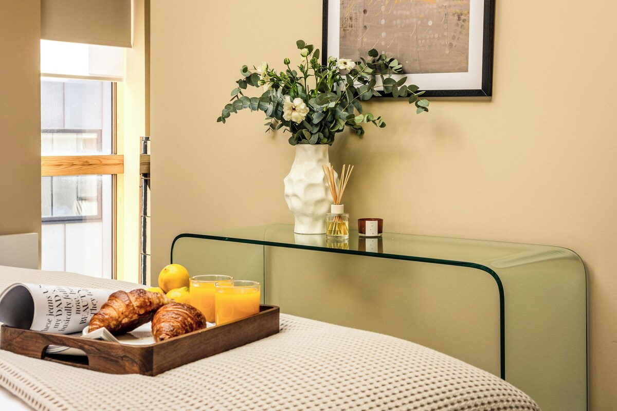 A neatly arranged breakfast tray with croissants and orange juice on a bed with a white textured blanket. A vase with flowers and a candle is on a glass table beside the bed.