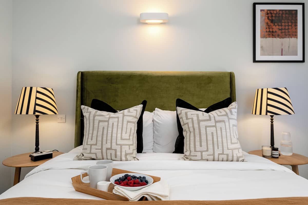 A neatly made bed with a green headboard, two bedside tables with lamps, and a tray with fruits and drinks.