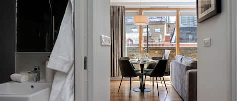 A modern bathroom with a sink, towel, and shower. It leads to a dining area with a table, chairs, and a view of the city.