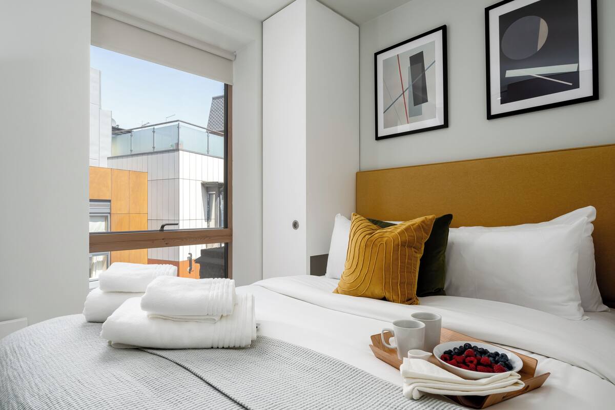 A hotel room with a large bed, white bedding, a wooden headboard, and a view of buildings through a window.