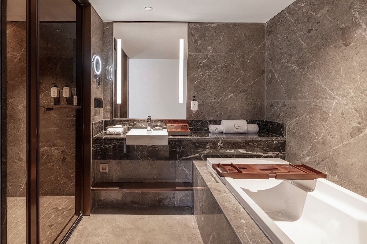 A modern bathroom with a large mirror, two sinks, a bathtub, and marble walls.