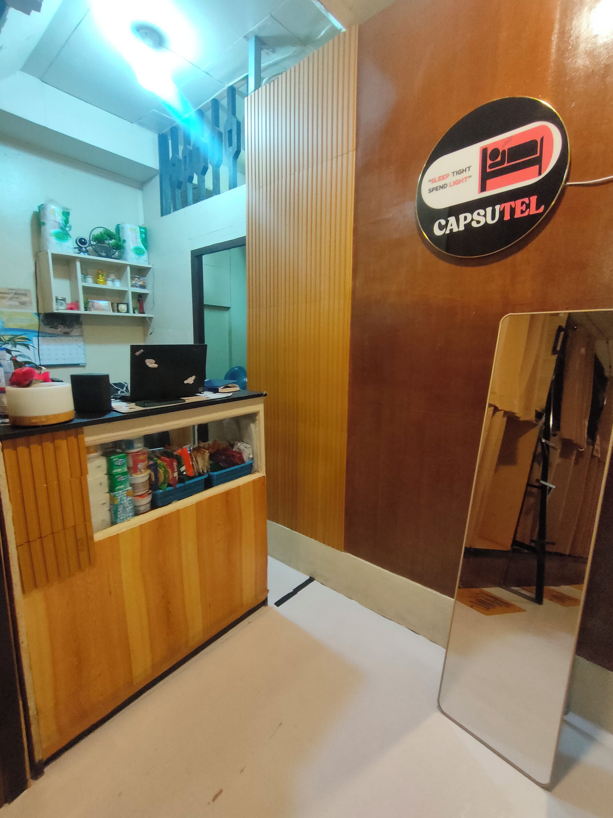 Un mostrador de recepción de hotel con una computadora, un espejo y un letrero que dice 'CAPSULE'.