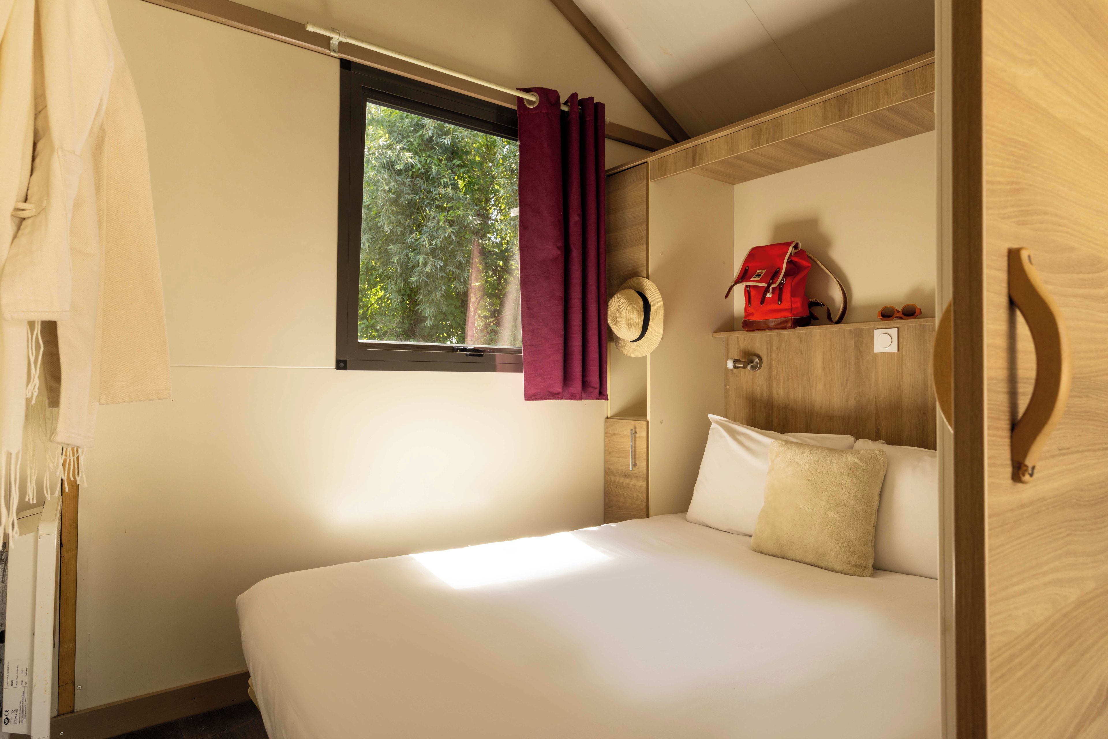 A single bed with a headboard, a window with a curtain, a small wooden cabinet, and a hat hanging on the wall.