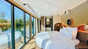 A modern bedroom with a large bed, a wooden headboard, a nightstand, a mirror, and a view of a lake through floor-to-ceiling windows. - Royale Escale Torcy (Torcy)