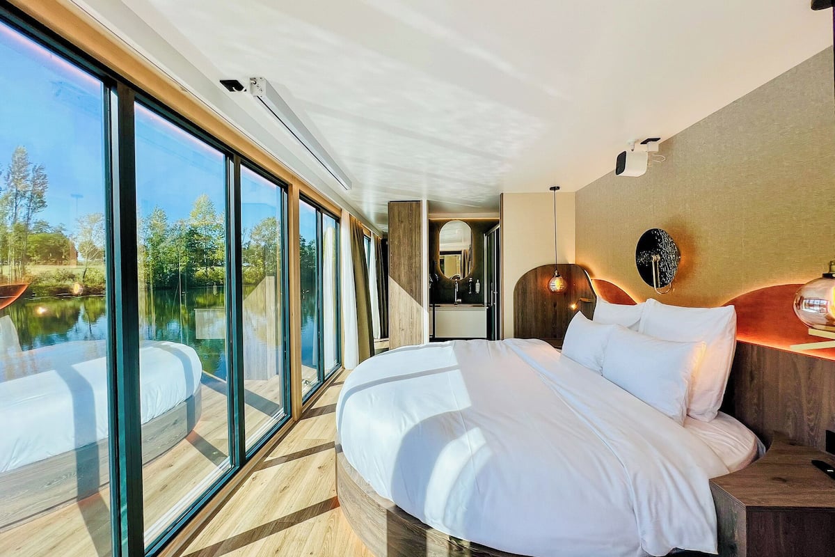 Una habitación de hotel moderna con una cama grande, cabecera de madera, un baño con espejo y vistas a un lago a través de ventanales de piso a techo.