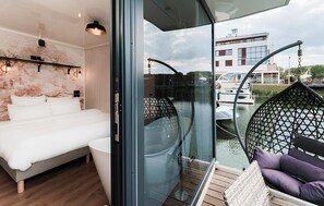 A modern bedroom with a bed, a glass-enclosed balcony with a view of a building and a boat.