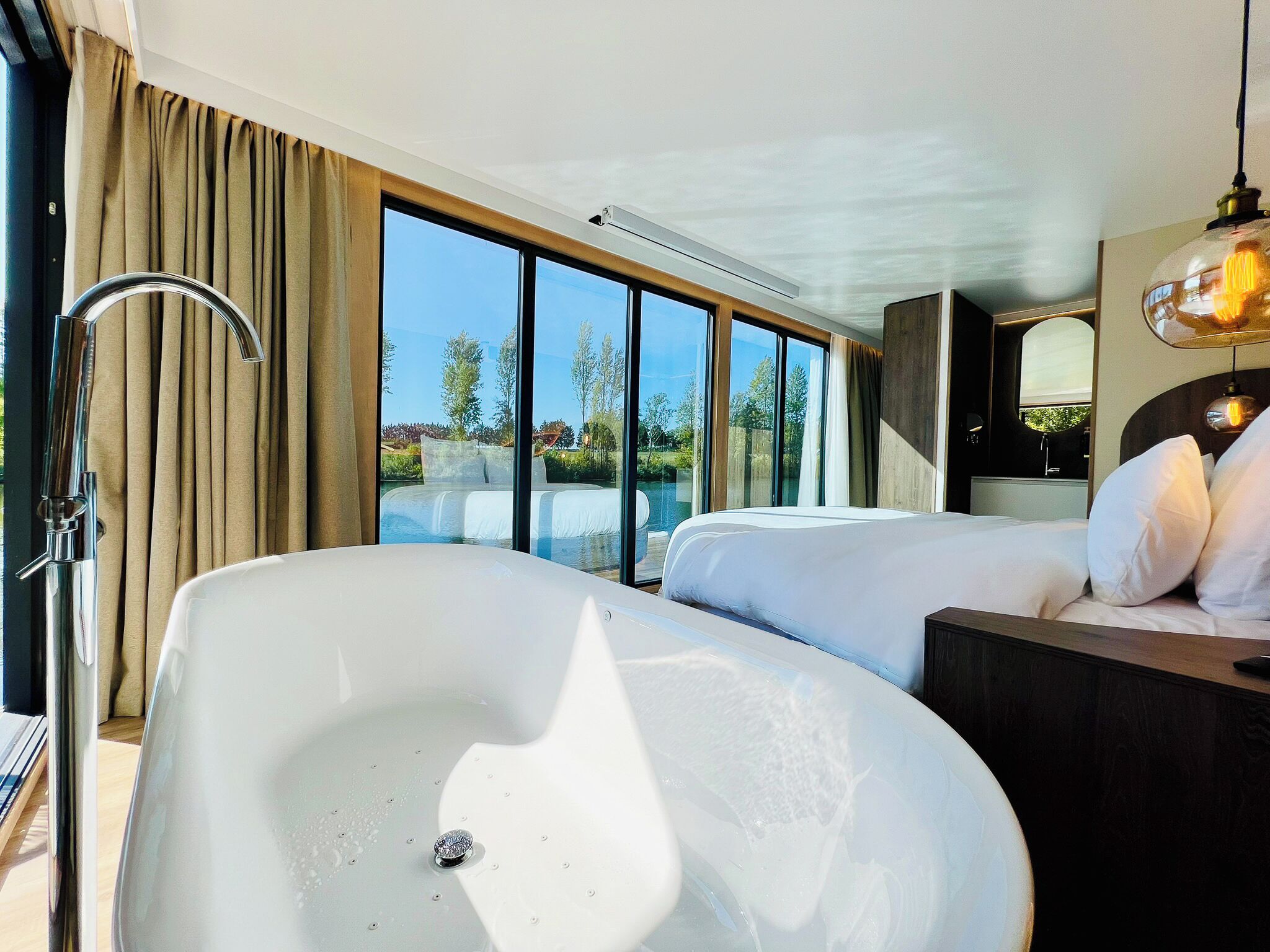 A modern hotel room with a large bathtub, a bed, a vanity with a mirror, and a view of a pool and trees.