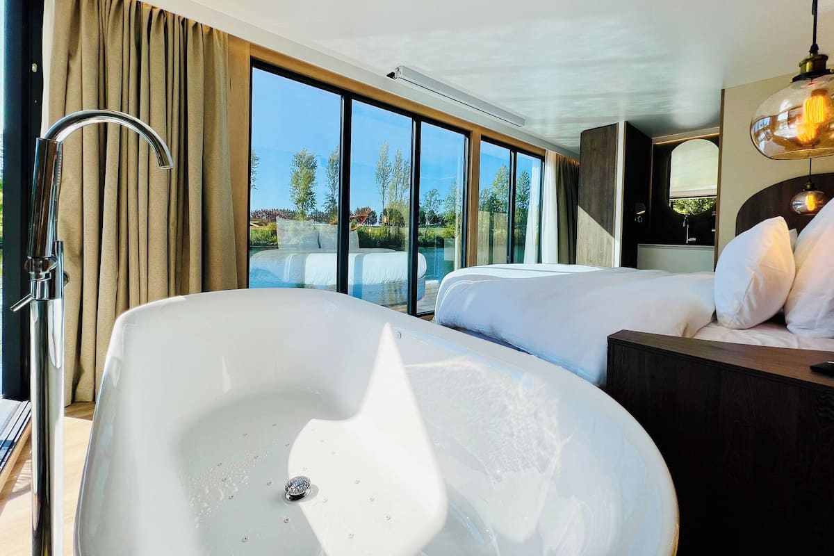 Habitación de hotel moderna con una bañera grande, una cama, un tocador con espejo y vistas a una piscina y árboles.