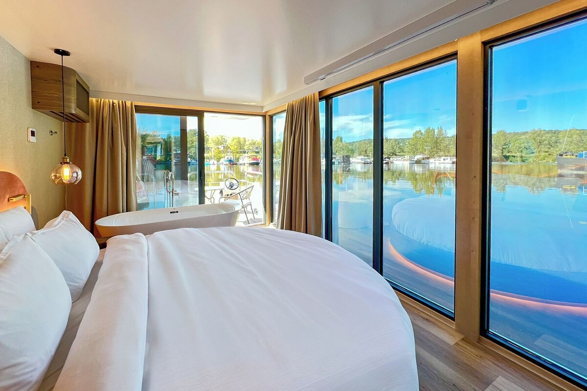 A hotel room with a large bed, a small round table, and a view of a body of water through large windows.