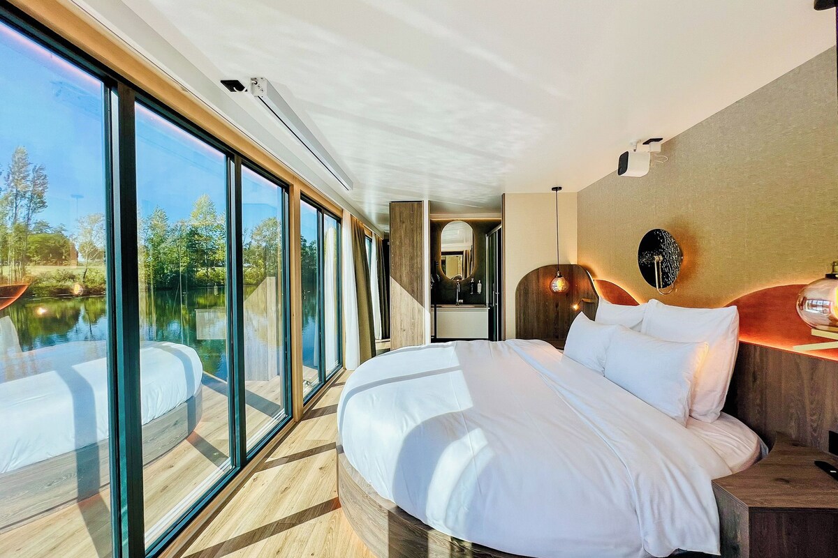 A modern bedroom with a large bed, a wooden headboard, a nightstand, a mirror, and a view of a lake through floor-to-ceiling windows.