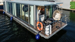 Une péniche flottante dotée d’un espace de vie entièrement vitré, d’une petite terrasse avec des sièges et d’une bouée de sauvetage.
