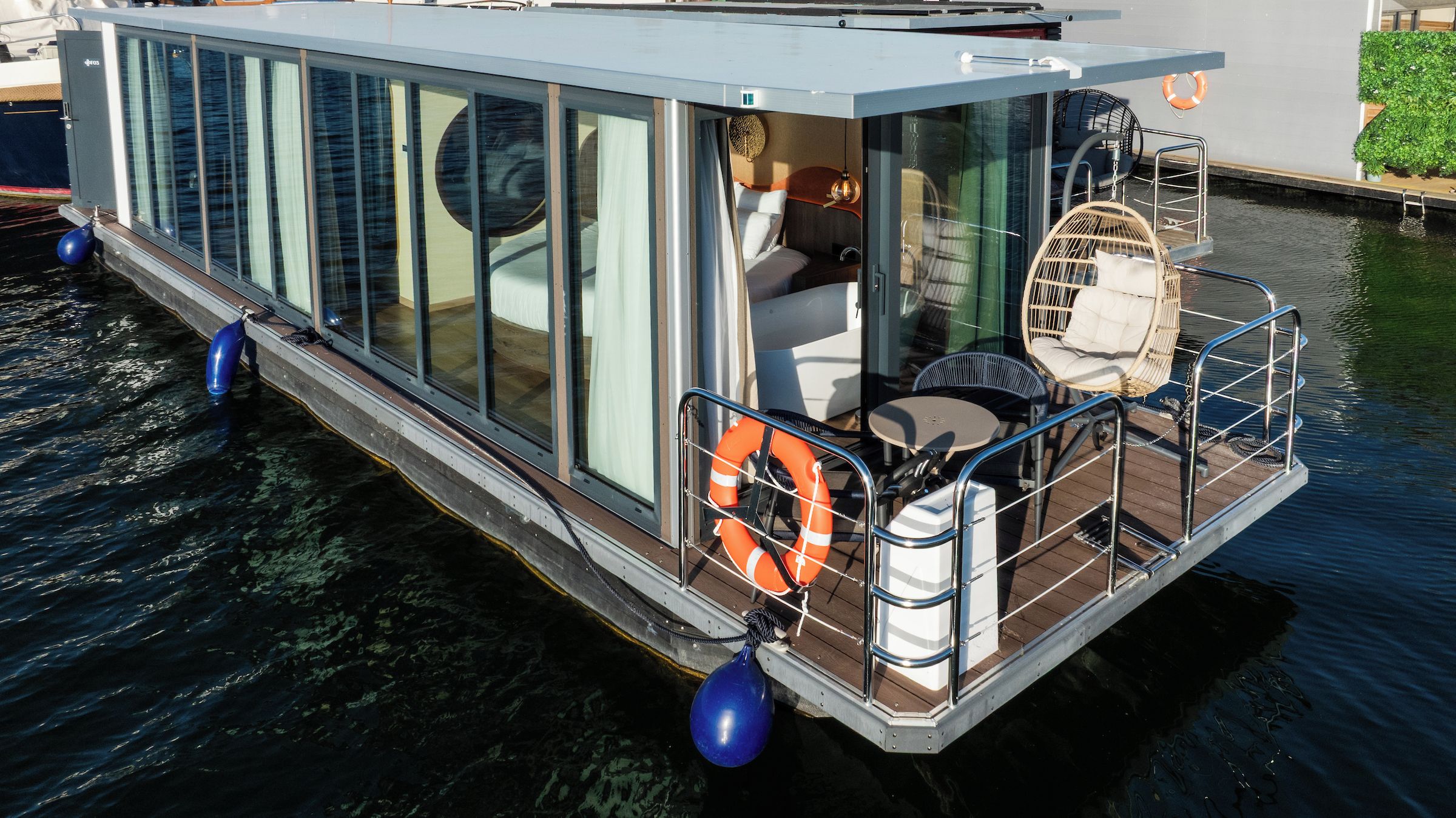 A modern floating home with a glass exterior, a small deck with seating, and a lifebuoy.