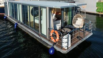 A modern floating home with a glass exterior, a small deck with seating, and a lifebuoy.