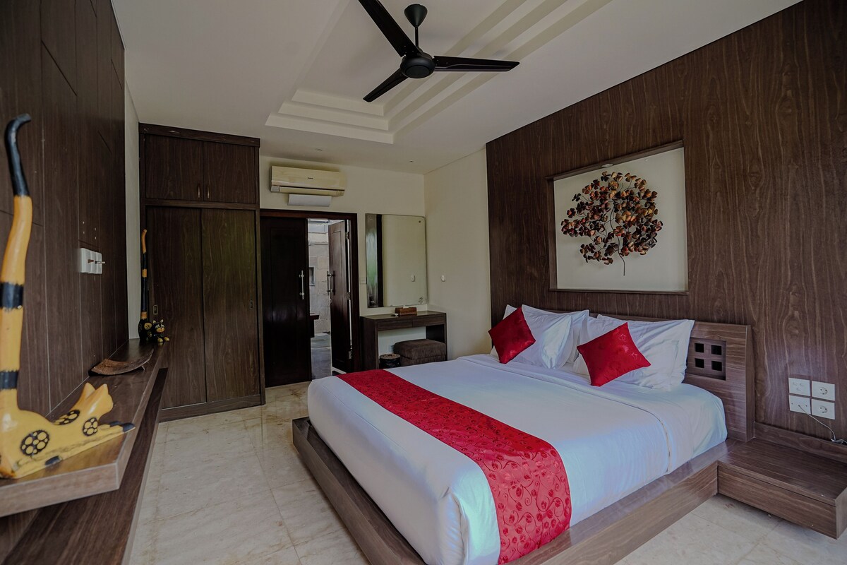 A hotel room with a large bed, wooden headboard, a ceiling fan, a wall-mounted artwork, and a wooden wardrobe.