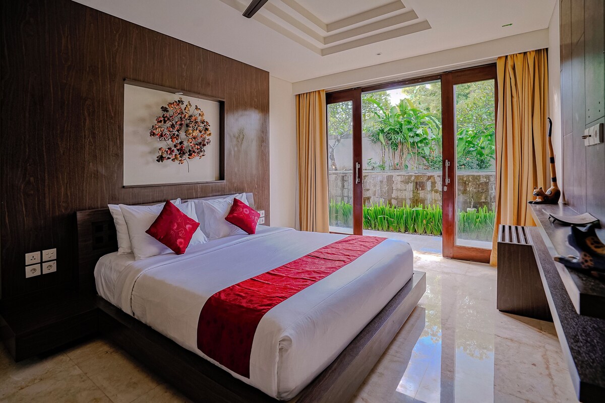 A modern hotel room with a large bed, a wooden headboard, a wall-mounted artwork, and a sliding glass door leading to a balcony with greenery.