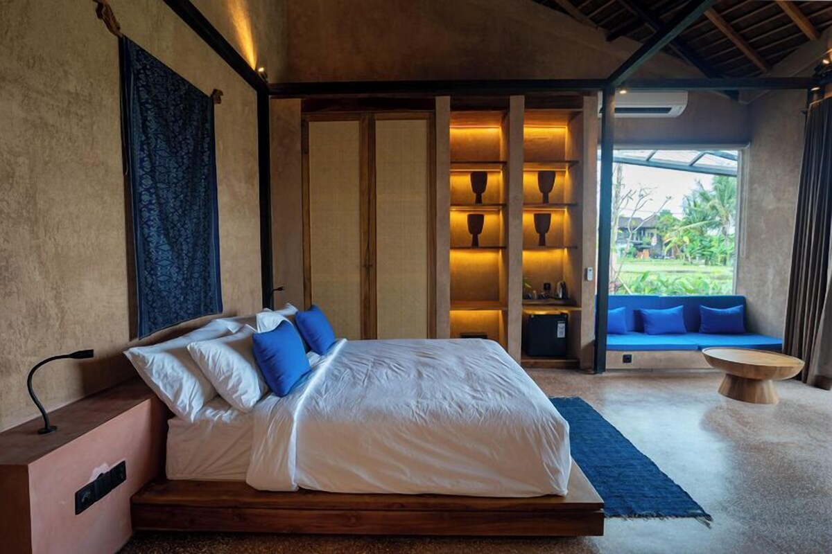 A bedroom with a bed, bedside table, and a view of a blue sofa and greenery outside.