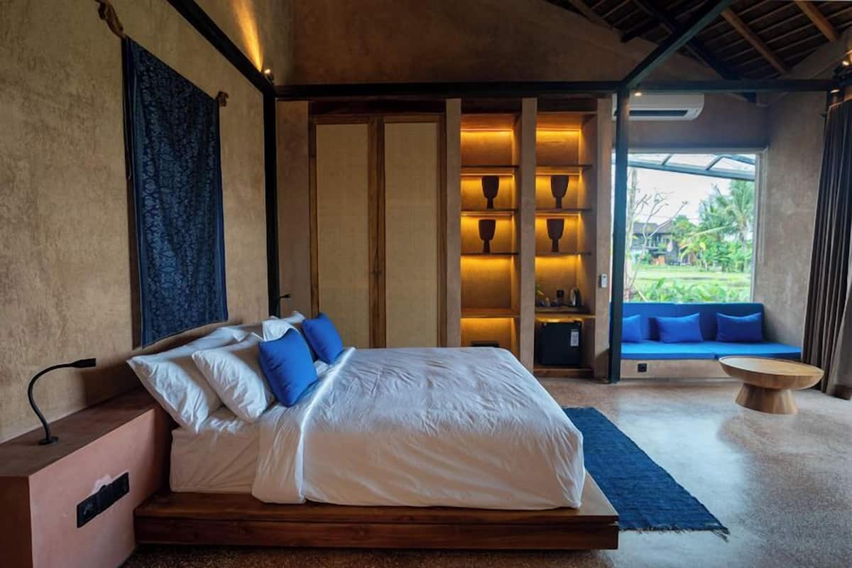 A bedroom with a bed, bedside table, and a view of a blue sofa and greenery outside.