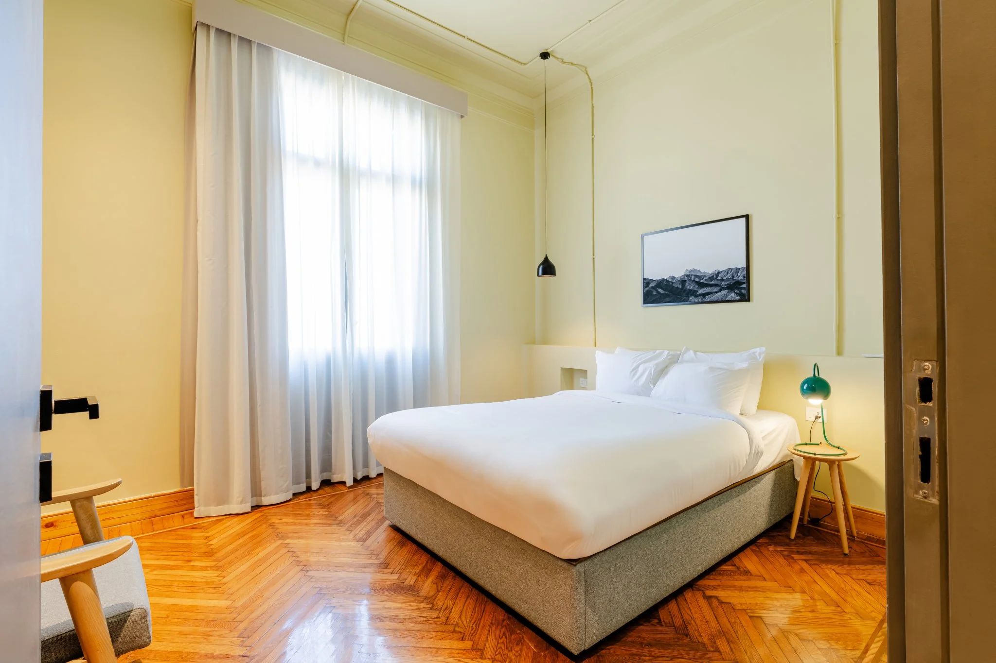 Un dormitorio ordenado con cama, mesita de noche, lámpara y ventana con cortinas.