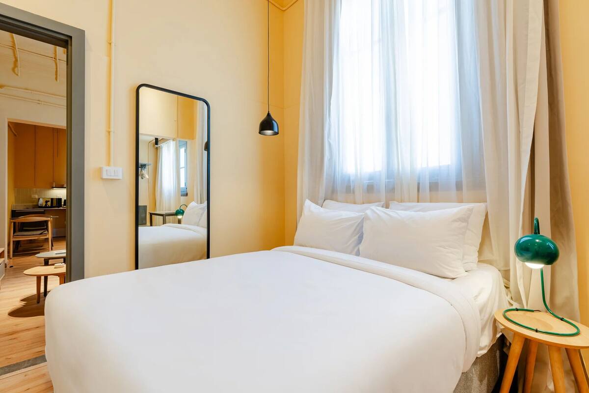 Ein Hotelzimmer mit Bett, Spiegel, Nachttischlampe und Blick ins Freie durch durchsichtige Vorhänge.