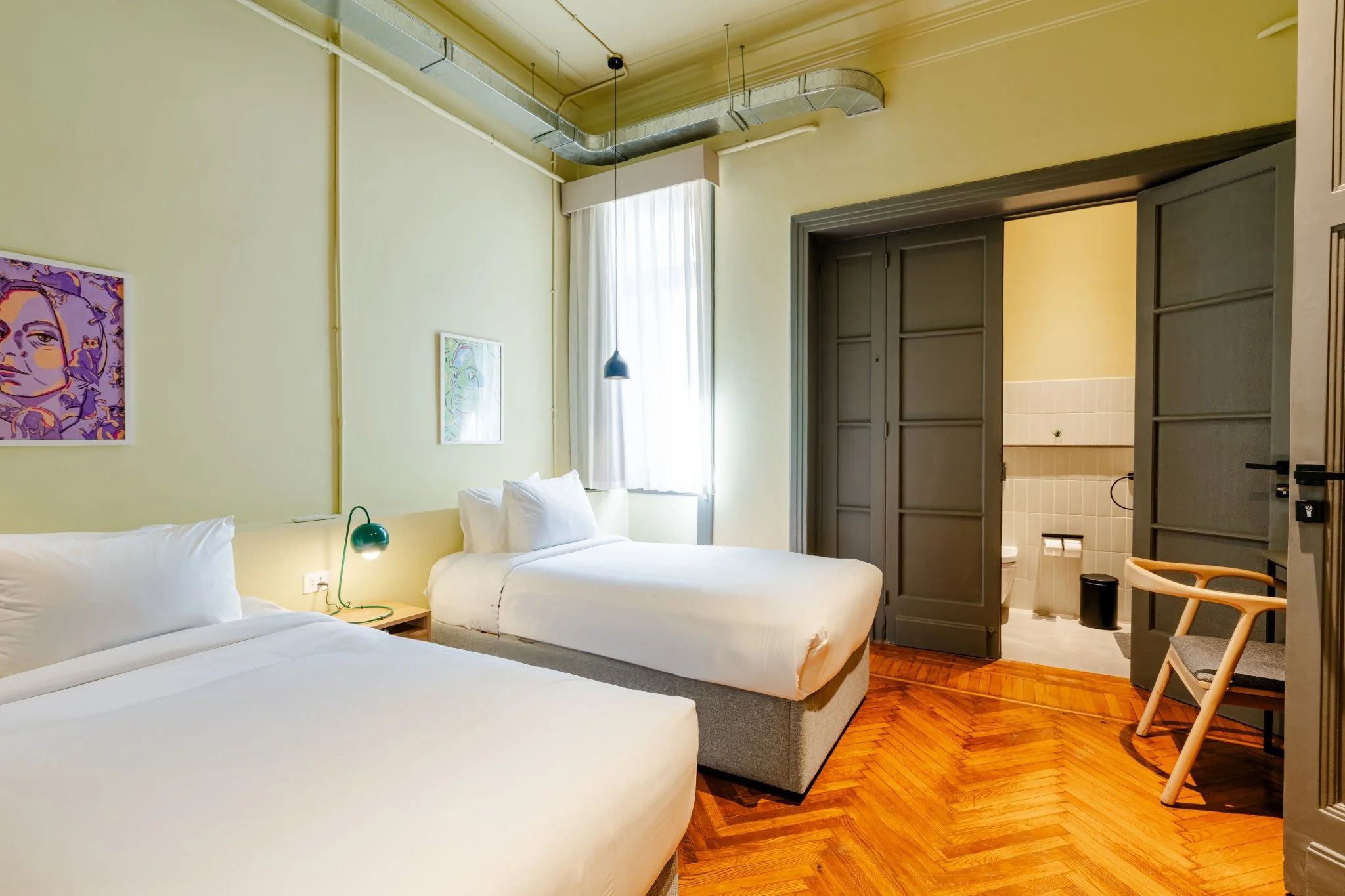 A hotel room with two beds, a wooden floor, a window with curtains, a door, and a small chair.