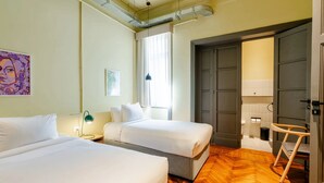 Une chambre d’hôtel avec deux lits, un plancher en bois, une fenêtre avec des rideaux, une porte et une petite chaise.