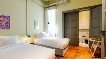 A hotel room with two beds, a wooden floor, a window with curtains, a door, and a small chair.