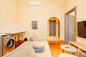 A compact living space with a sofa, a washing machine, a small table, a TV, and an arched doorway. - Studio Apartments Downtown Alexandria Menascehouse (Alexandria)