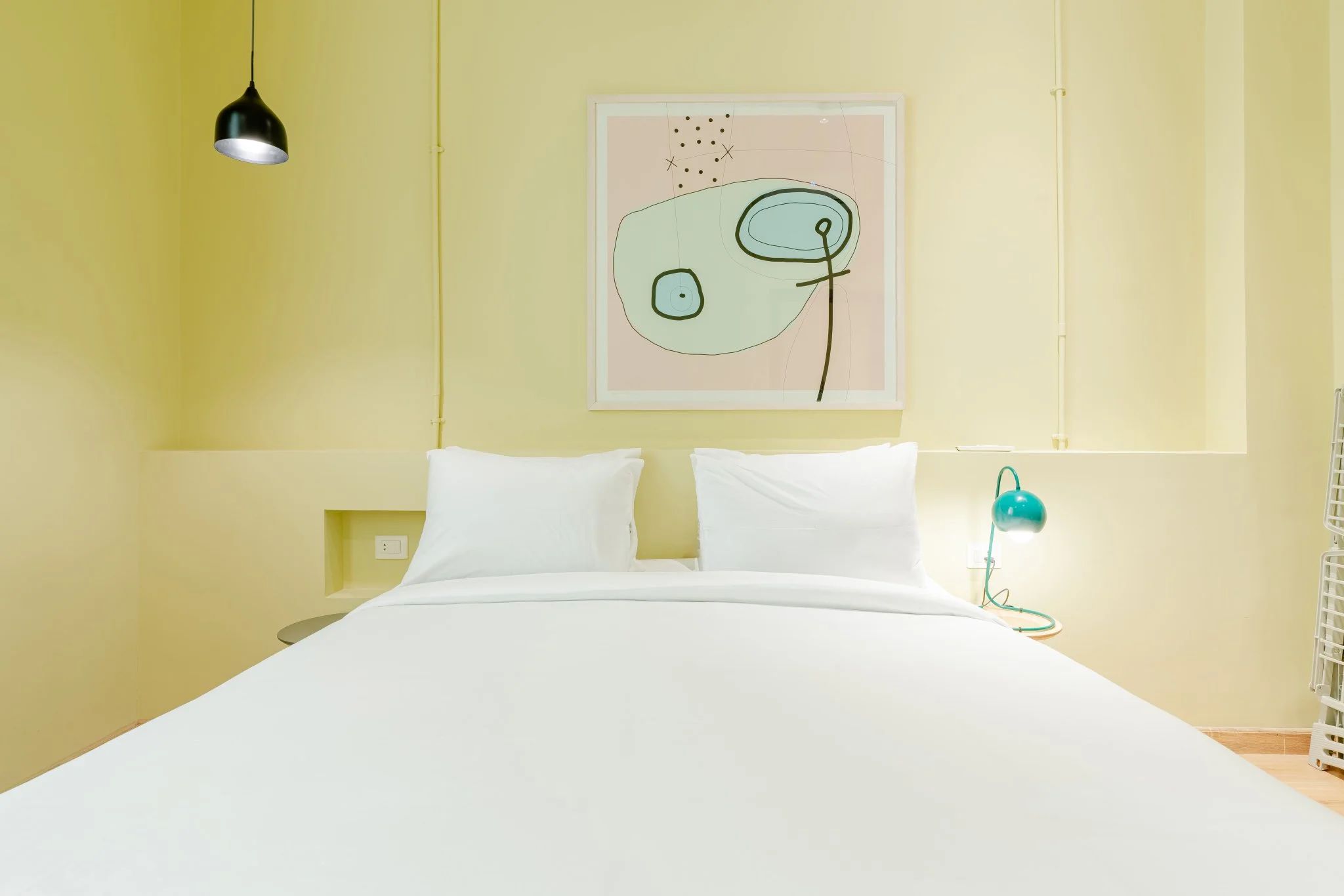 Un lit avec une literie blanche, une suspension noire, une lampe de bureau turquoise et une œuvre d’art abstraite accrochée au mur.