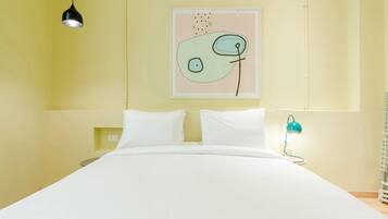Una cama con ropa de cama blanca, una lámpara colgante negra, una lámpara de escritorio azul verdoso y una obra de arte abstracto en la pared.