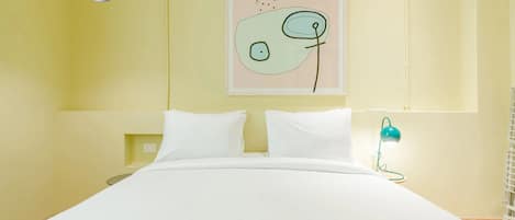 A bed with white bedding, a black pendant light, a teal desk lamp, and a wall-mounted abstract art piece.