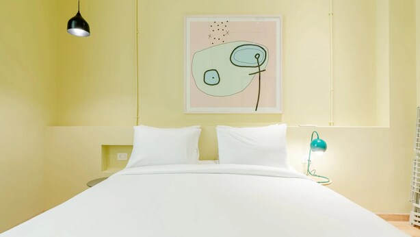 A bed with white bedding, a black pendant light, a teal desk lamp, and a wall-mounted abstract art piece. - Studio Apartments Downtown Alexandria Menascehouse (Alexandria)
