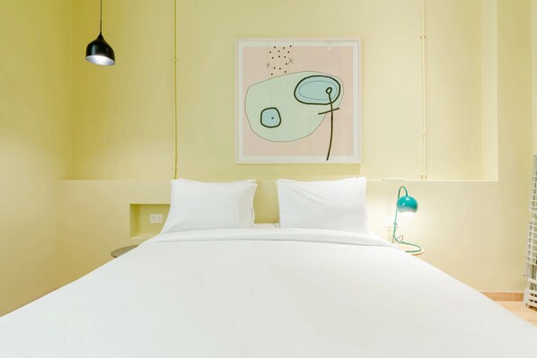 A bed with white bedding, a black pendant light, a teal desk lamp, and a wall-mounted abstract art piece. - Studio Apartments Downtown Alexandria Menascehouse (Alexandria)