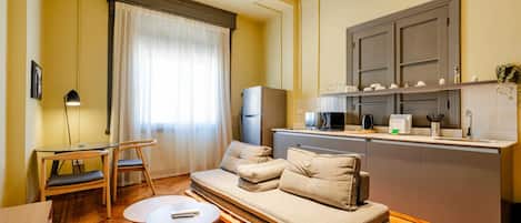 Un espacio habitable compacto con sofá, una pequeña mesa de comedor, kitchenette y una ventana con cortinas.
