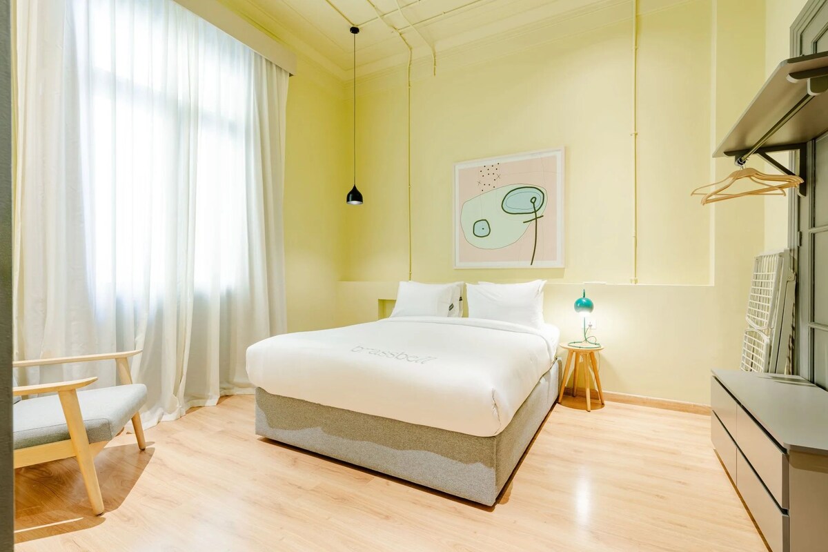 A bedroom with a bed, a chair, a bedside table with a lamp, a wall art piece, and a window with curtains.