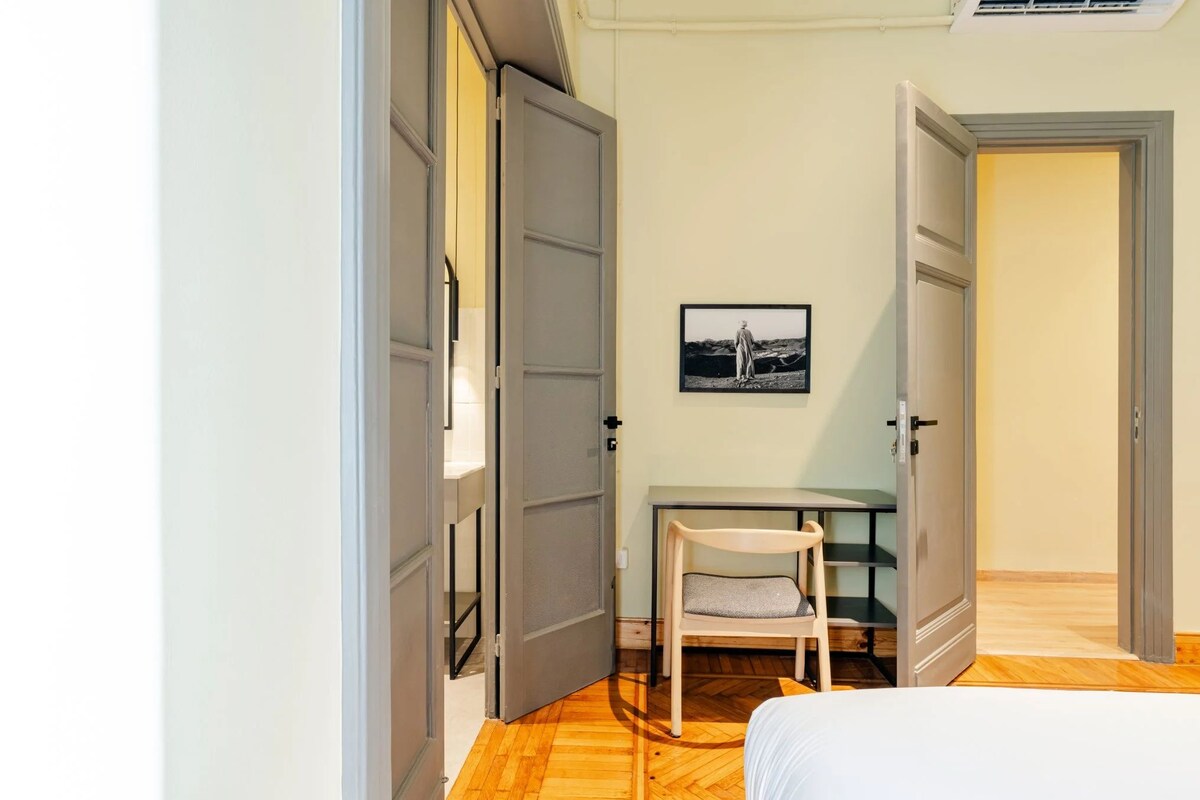 A bedroom with a bed, a desk with a chair, and a door leading to another room.