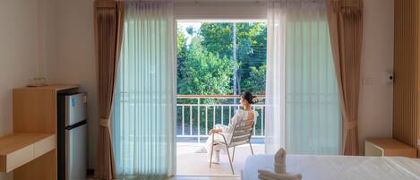 A hotel room with a bed, a refrigerator, a window with curtains, and a view of greenery outside.