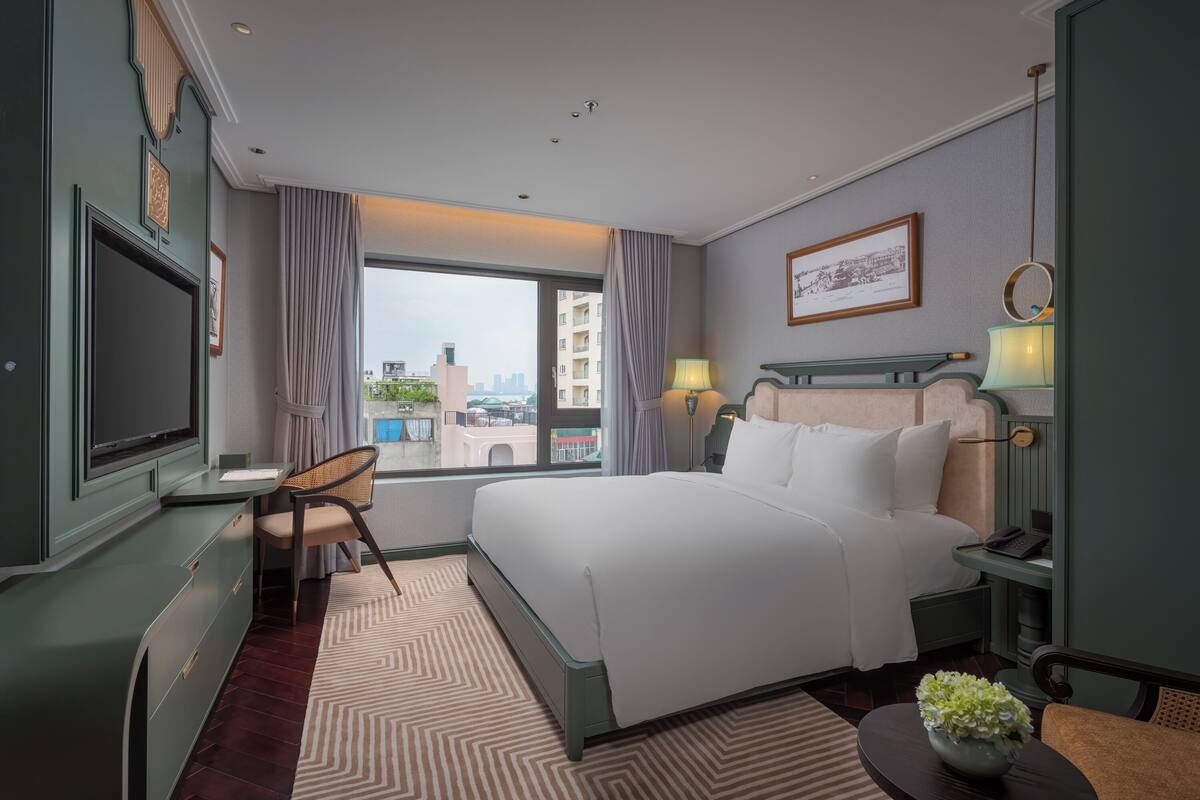 A hotel room with a bed, bedside table, lamp, chair, TV, and a view of the cityscape through the window.