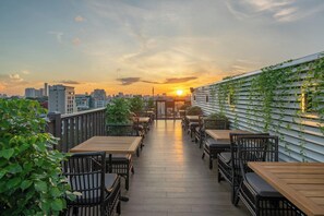 Una terraza en la azotea con mesas y sillas de madera, un horizonte urbano al fondo y una puesta de sol.