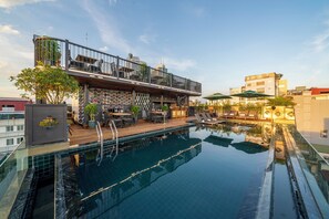 Un área de piscina en la azotea con bar y asientos tipo lounge, rodeada de edificios de la ciudad.