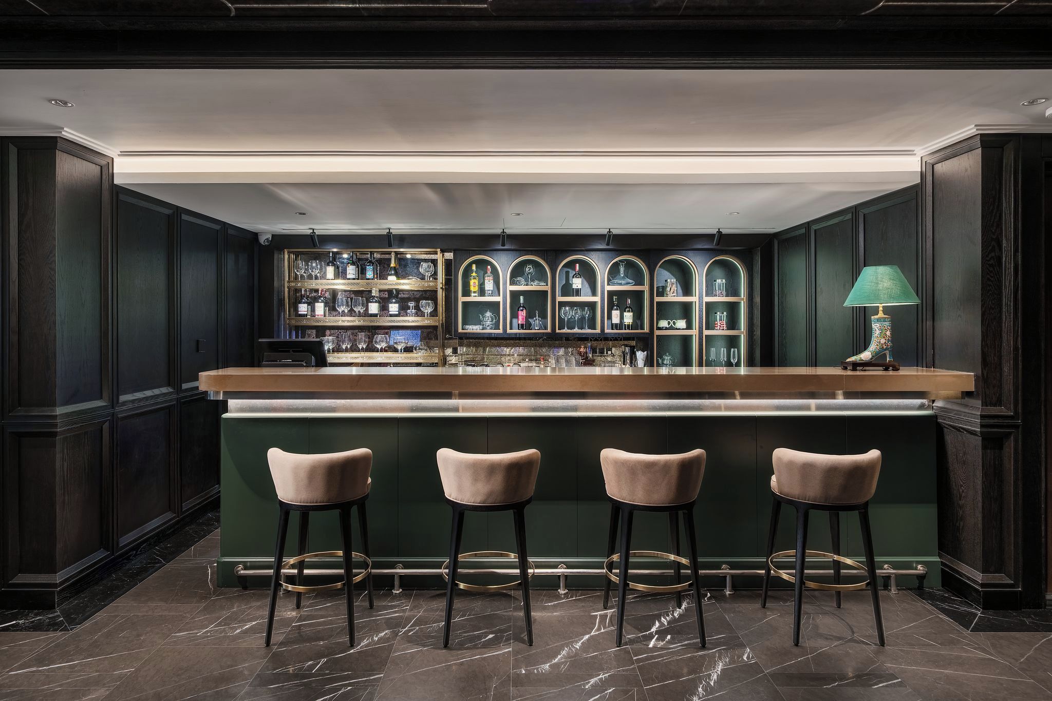A bar area with a counter, three cushioned bar stools, and shelves with bottles and glasses.