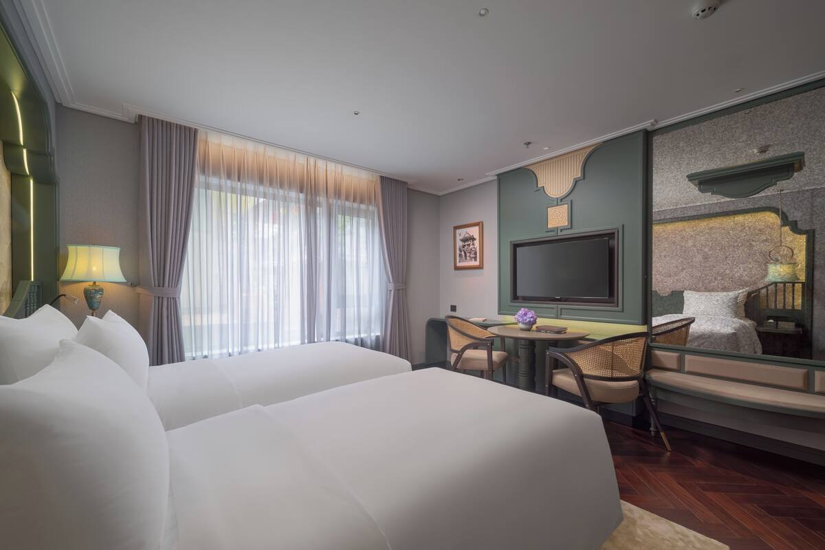 A hotel room with two beds, a TV, a desk with chairs, and a large window with curtains.