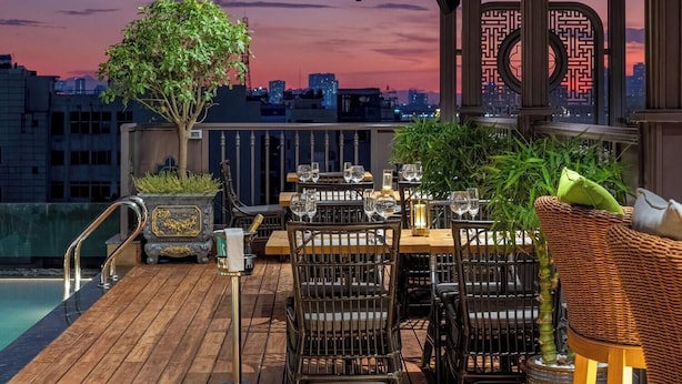Un espacio exterior para comer con mesas y sillas de madera, una piscina y vistas a la silueta de la ciudad al atardecer.