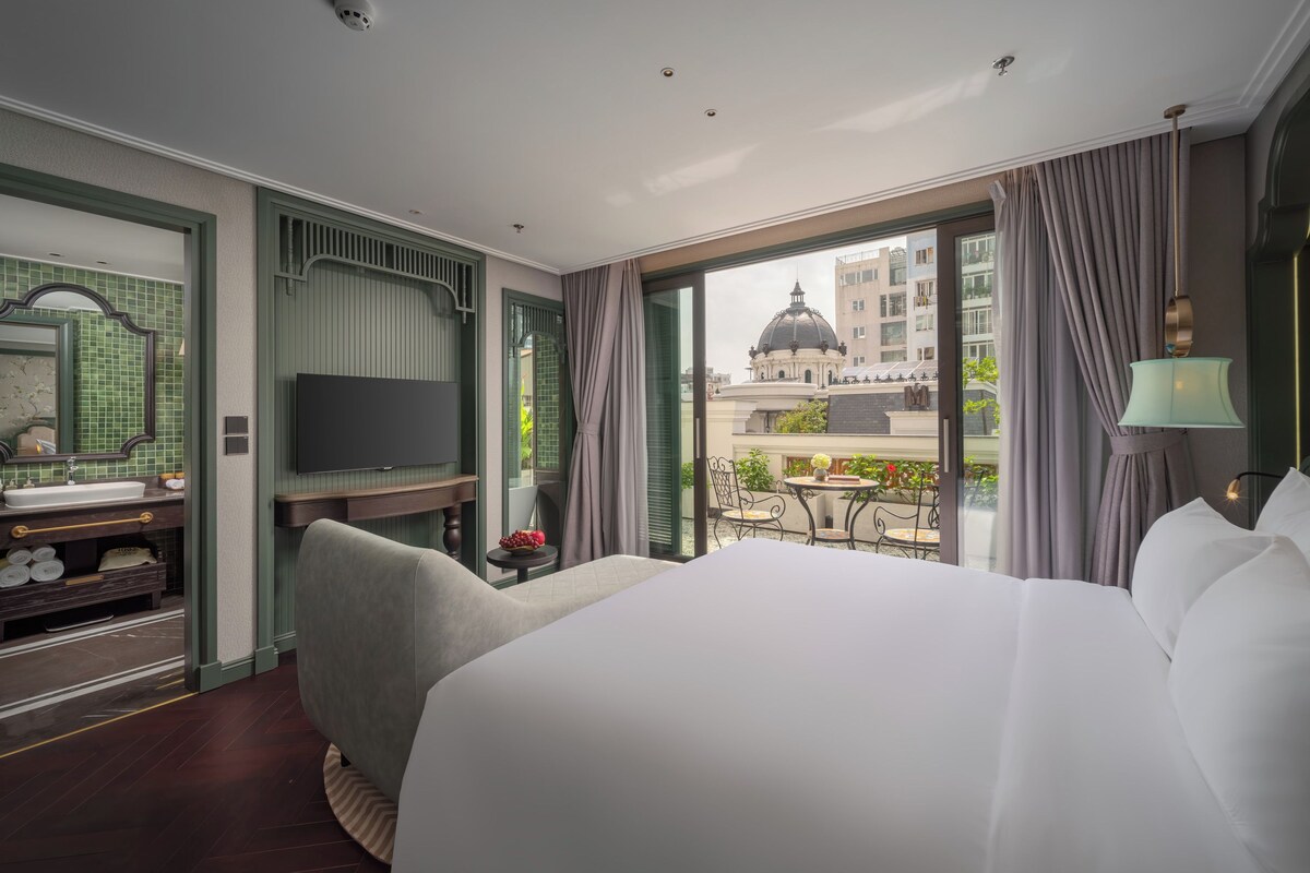 A hotel room with a large bed, a TV, a balcony with a view of buildings, and a small table with flowers.