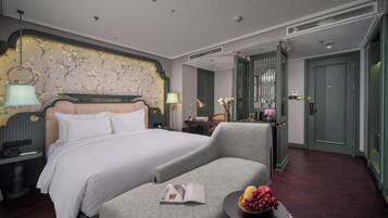 A hotel room with a bed, a reading chair, a small table with fruit, a desk with a lamp, and a decorative headboard.