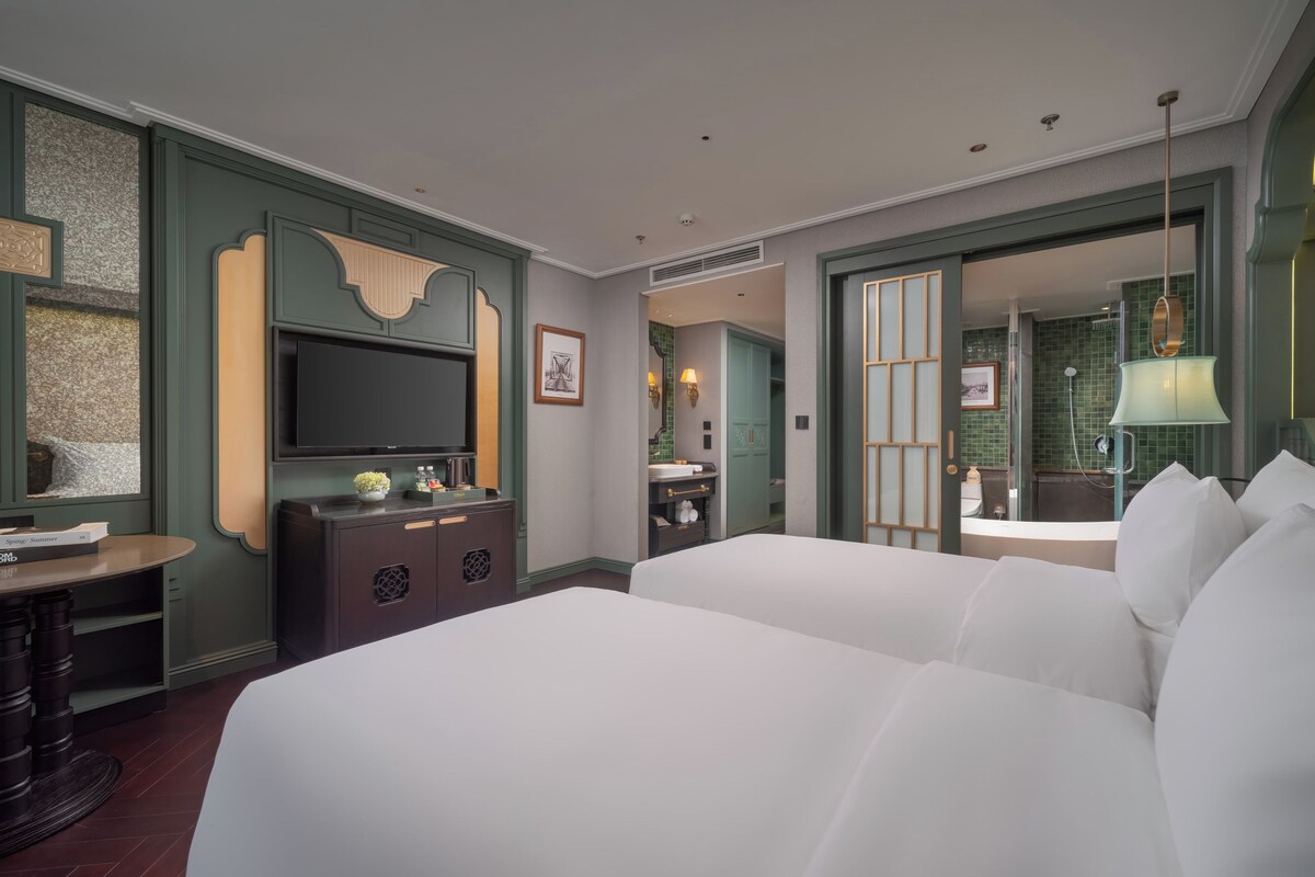 A hotel room with a large bed, a TV, a desk with a chair, a dresser, and a bathroom visible through an open door.