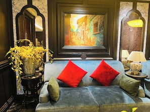 Una habitación con un sofá azul, cojines rojos, un jarrón con flores amarillas, un cuadro de una escena callejera y una lámpara sobre una mesita.