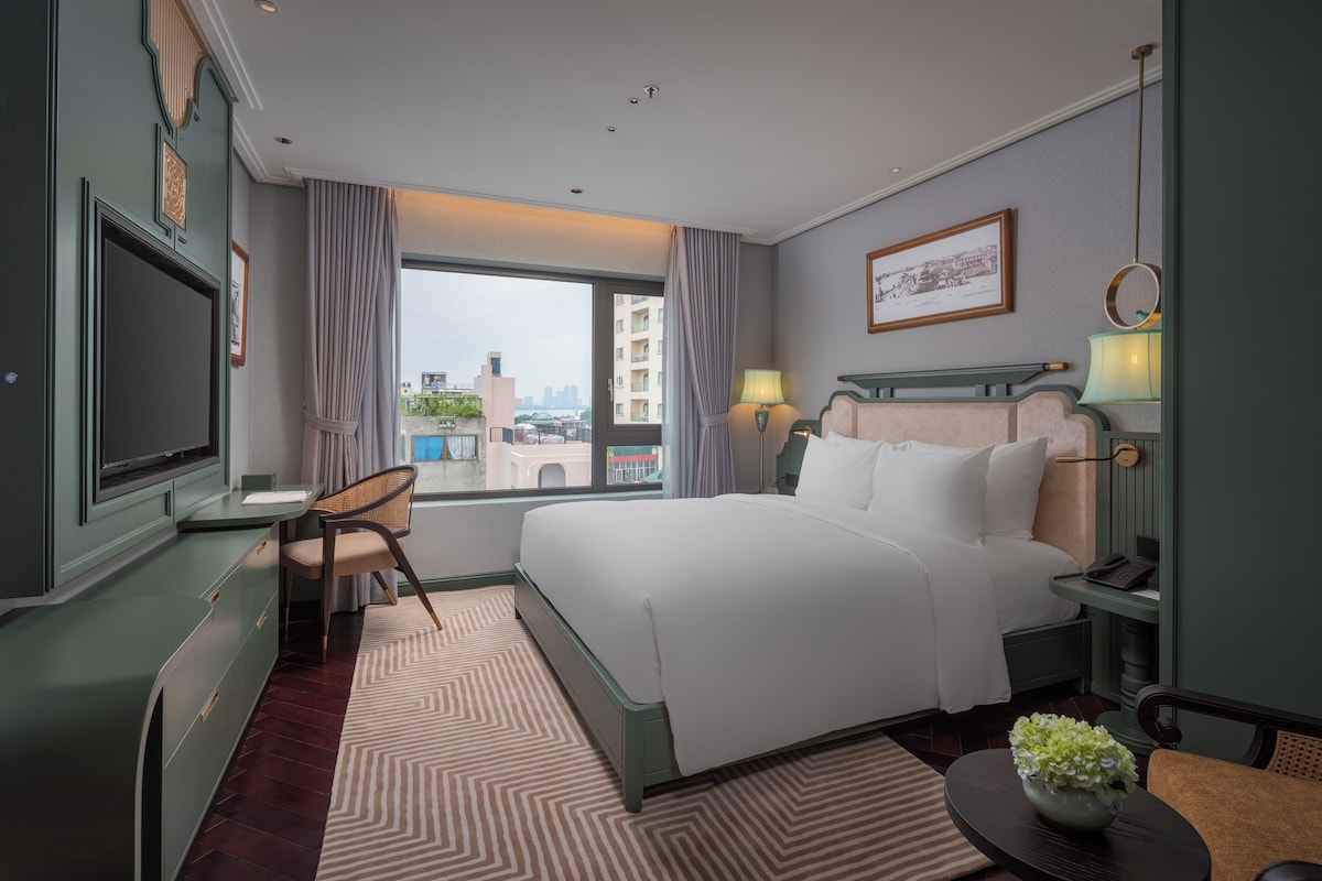 A hotel room with a bed, a desk, a chair, a TV, a window with a view of buildings, and a lamp.