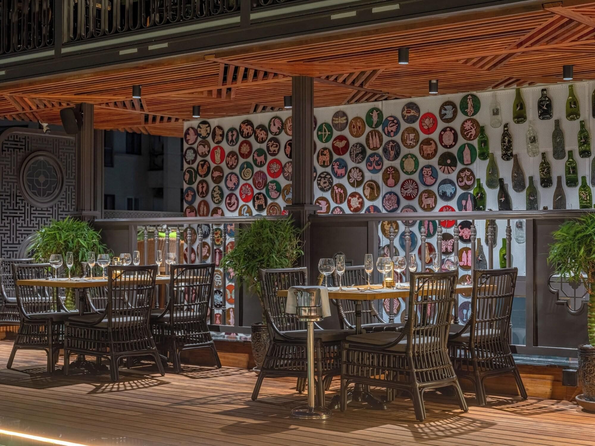 An outdoor dining area with wicker chairs and tables, a wooden ceiling, and a wall decorated with circular art pieces.