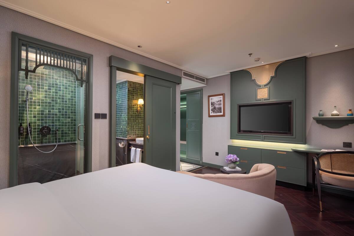 A modern hotel room with a large bed, a desk with a flat-screen TV, a seating area, and a bathroom with a green-tiled shower area.